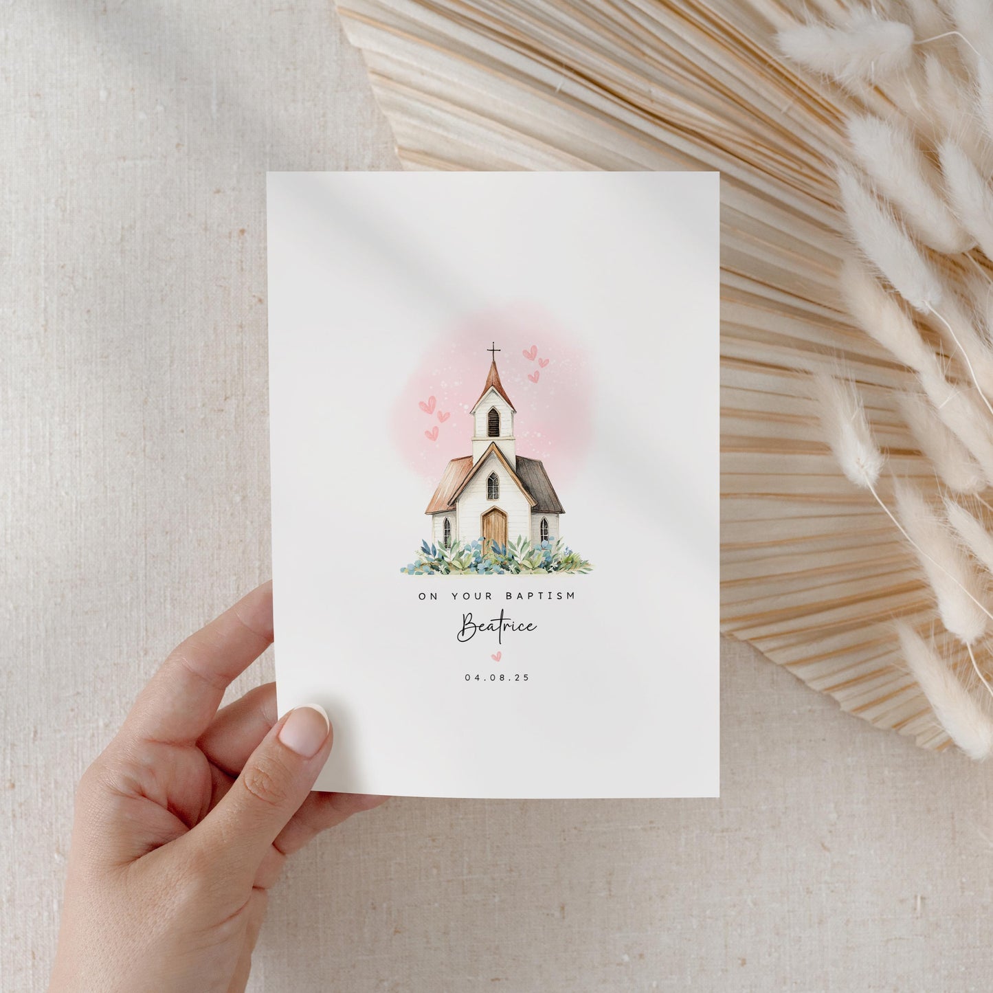 A hand holds a white card featuring a soft illustration of a church with a pink sky and small hearts. The text below reads “On Your Baptism, Beatrice, 04.08.25.” The card rests against a soft neutral background with pampas grass for decoration.