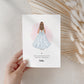 A hand holds a card featuring a girl in a white dress with a large bow on the back. The card says “Special Blessings on Your First Holy Communion” with the name “Hollie” below. The background includes natural dried wheat decor.