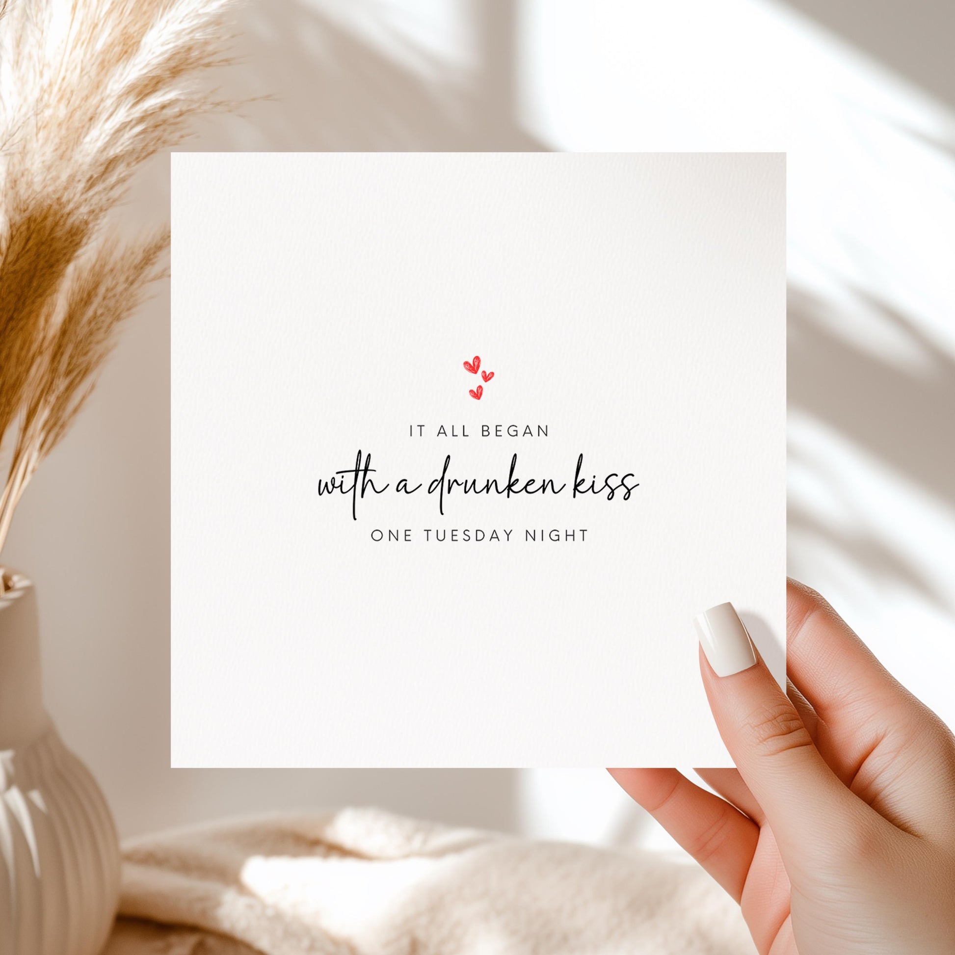 White card with “It all began with a drunken kiss one Tuesday night” design in black script with red hearts.